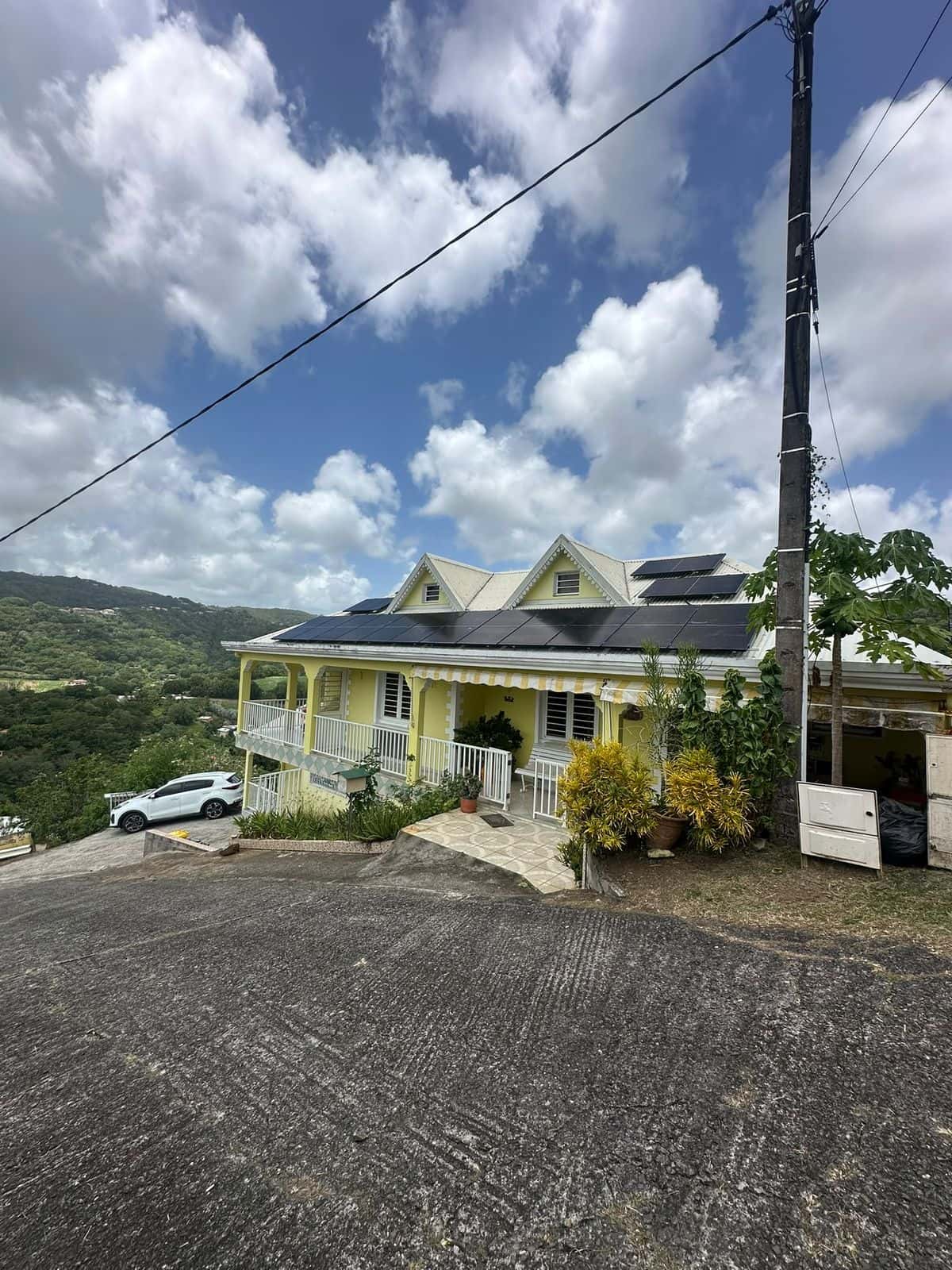 Maison créole jaune à balcons, panneaux solaires noirs sur le toit, mornes en fond, Martinique