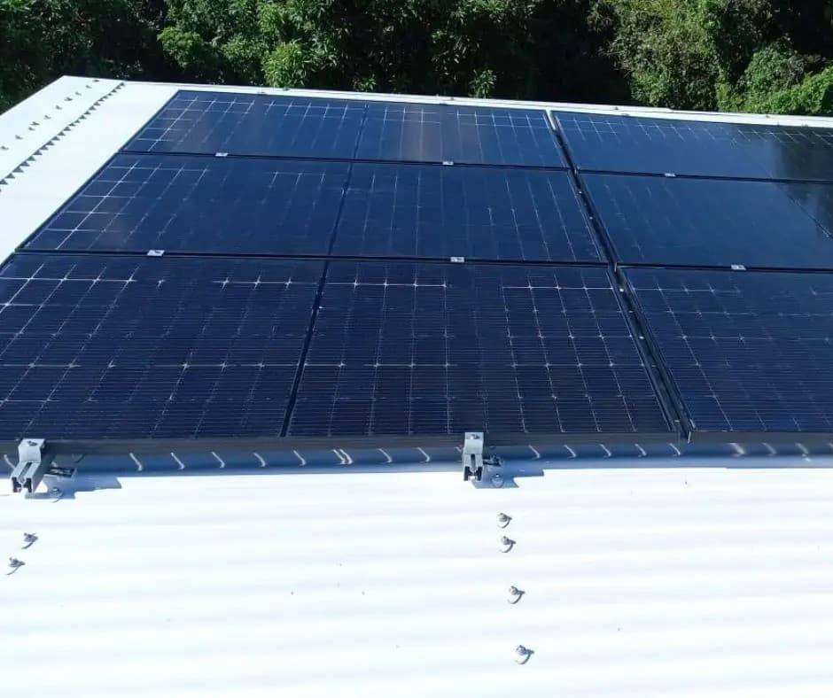 Deux rangées de panneaux solaires noirs sur une toiture en tôle blanche, végétation en arrière-plan